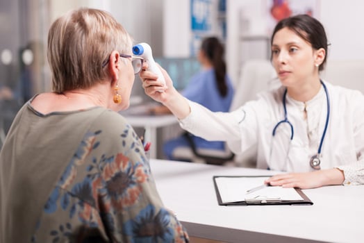 Young doctor using infrared thermometer to take senior woman temperature in hospital office. Medic wearing white coat and stethoscope and nurse working on computer in the background. photo