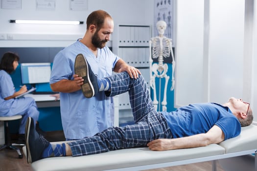 Physiotherapist performs leg exercise to stretch muscles and crack bones of elderly man in hospital cabinet. Retired patient receives kinesiology treatment for knee injury with assistance from nurse. photo