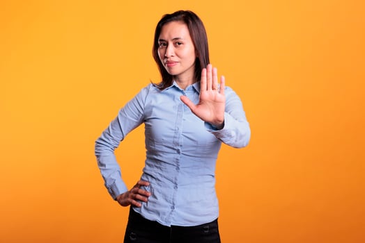 Serious filipino woman doing rejection gesture with hand in front of camera, posing in studio over yellow background. Negative model showing stop gesture, being in denial and expressing refusal. photo