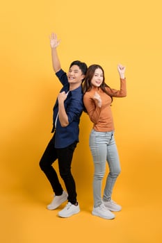 Full body view of cheerful Asian couple standing back to back smiling and clenching fists in yellow isolated studio background. photo