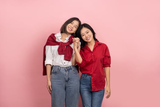 Cheerful young Asian lesbian couple in shirts and jeans stands holding hands to express their love on a pink background. photo