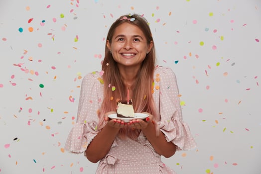 Portrait of cheerful young lovely woman with light brown long hair posing in multicolored confetti with piece of cake in raised hands, looking upwards and smiling pleasantly photo