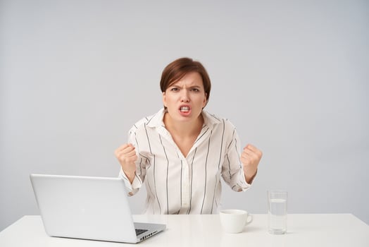 Furious young brown haired female with short trendy haircut clenching her fists and frowning face while looking violently at camera, posing over white background in striped shirt photo