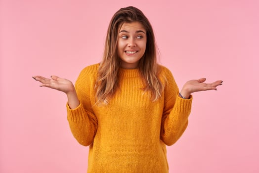 Bewildered young brown haired woman with natural makeup biting confusedly her underlip while standing over pink background with raised palms, dressed in casual wear photo