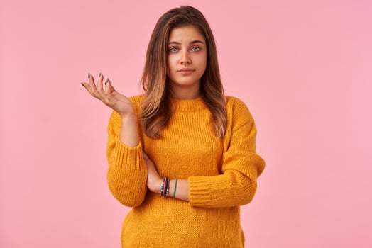 Displeased young brown haired lady with natural makeup raising hand perplexedly and looking at camera with pout, keeping lips folded while standing over pink background photo