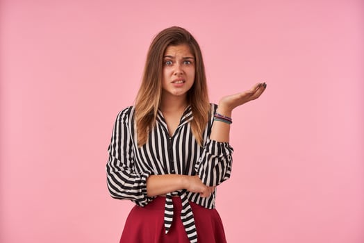 Displeased young pretty brunette female with natural makeup raising confusedly palm and looking at camera with pout, wearing striped shirt and red skirt over pink background photo