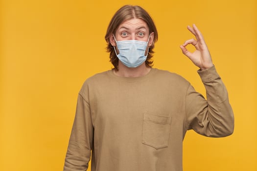 Happy looking male, positive bearded guy with blond hairstyle. Wearing beige sweater and medical protective face mask. Showing okay sign. Watching at the camera isolated over yellow background photo