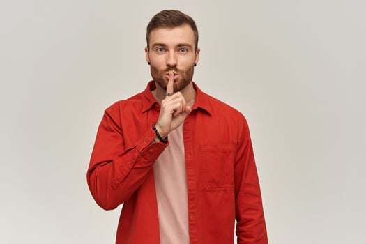 Happy attractive young bearded man in red shirt showing silence gesture and looking at camera over white background photo