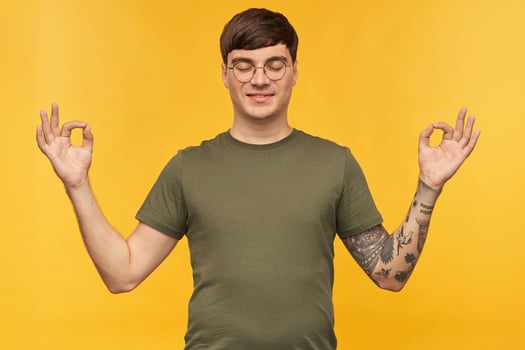 Smiling young male, shows mudra sign while meditating, wears green t-shirt and round glasses. Isolated over yellow studio background. photo