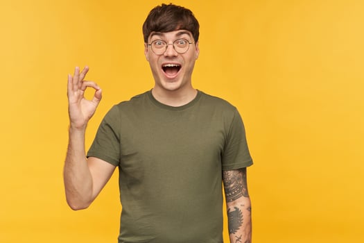 Indoor portrait of young man, shows ok sign with his hand, smiles broadly and looking directly into camera with happy facial expression. Isolated over yellow background. photo