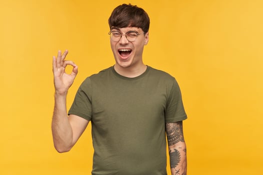 Indoor portrait of young man, shows ok sign with his hand, smiles broadly and wink with happy facial expression. Isolated over yellow background. photo