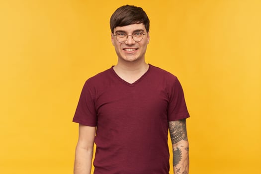 Indoor shot of young male , wears dark red t-shirt, round glasses and tattooed hand, looks directly into camera with broadly smile. Isolated over yellow background. photo