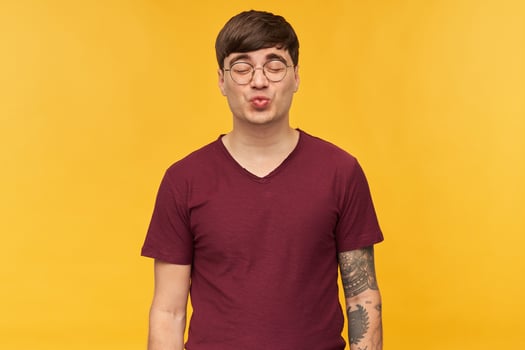 Young flirty male, wears dark red t-shirt and round glasses, keeps his eyes closed while kissing. Isolated over yellow background. photo