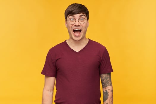 Indoor shot of amazed, surprised young male student, smiles broadly and keeps his eye widely opened with happy facial expression. Isolated over yellow background. photo