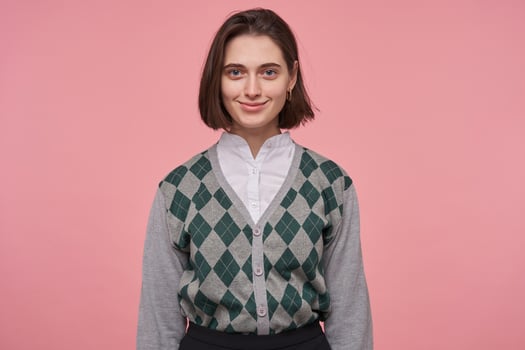 indoor shot of young brunette female wears stylish gray pullover smiling while starring into camera. Isolated over pink background. photo