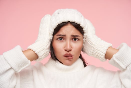 Bewildered young green-eyed brunette female with loose hair keeping raised hands on her head while looking confusedly at camera, standing over pink background photo