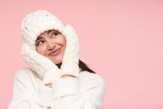 Pleased young attractive brown haired lady in white knitted hat keeping raised palms on her cheeks while looking gladly upwards with nice smile, posing over pink background photo