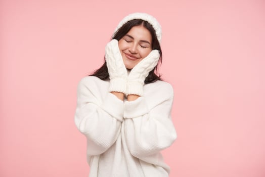 Pleasant looking young cheerful brown haired woman with casual hairstyle smiling gladly with closed eyes and holding palms on her cheeks, isolated over pink background photo