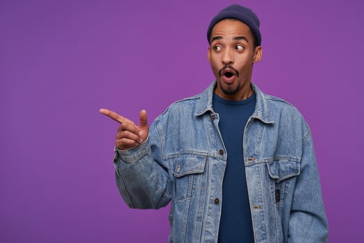 Surprised young brown-eyed dark skinned bearded man dressed in casual clothes showing amazedly aside with forefinger and keeping mouth opened, standing over purple background photo