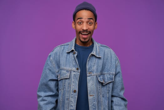 Open-eyed young beautiful dark haired bearded male in blue cap, pullover and jeans coat raising amazedly eyebrows while looking joyfully at camera, standing over purple background photo