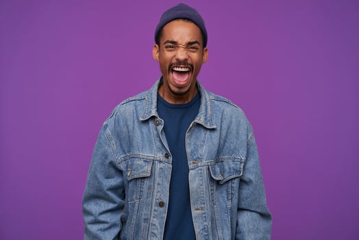 Indoor photo of young angry dark skinned brunette male with beard frowning his face while screaming fiercely with wide mouth opened, isolated over purple background