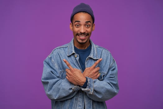 Indoor photo of cheerful young handsome dark skinned bearded man smiling widely while showing with forefingers in different directions, standing over purple background