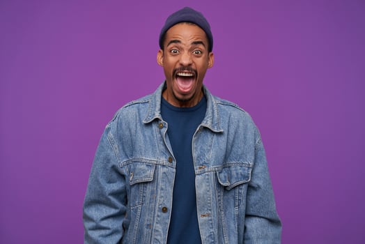 Furious young brown-eyed bearded man with dark skin shouting angrily and wrinkling forehead while looking heatedly at camera, wearing blue cap, pullover and jeans coat over purple background photo