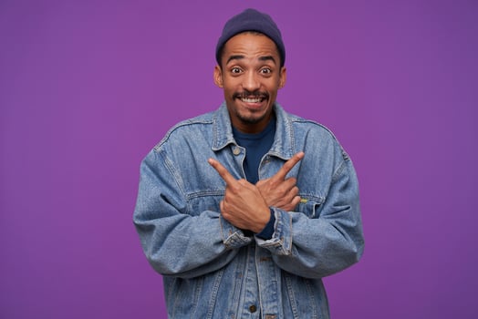 Emotional young lovely bearded brunette male with dark skin rounding his brown eyes while looking joyfully at camera and smiling broadly, standing over purple background photo