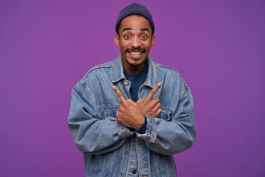 Overjoyed young brown-eyed dark skinned bearded male looking amazedly at camera with wide smile, showing in different sides with raised index fingers, isolated over purple background photo