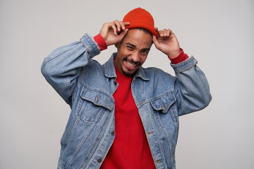 Cheerful young pretty dark skinned brunette male with beard holding his red cap with raised hands while standing over white background, being in nice mood and smiling happily photo