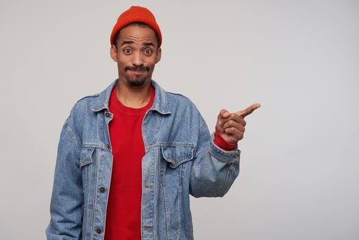 Confused young brown-eyed bearded dark haired male raising surprisedly eyebrows and puffing out cheeks while showing aside with index forefinger, isolated over white background photo