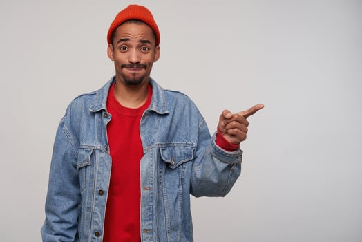 Studio photo of young dark skinned brunette man with beard getting air in his mouth and looking confusedly at camera, keeping index finger raised while posing over white background