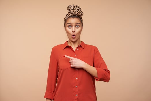 Bemused young lovely brown haired lady with headband rounding surprisedly her green eyes while looking amazedly at camera and pointing aside with forefinger, isolated over beige background photo
