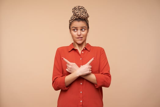 Bewildered young pretty brunette woman with headband biting worringly underlip while showing in different directions with index fingers, posing over beige background photo