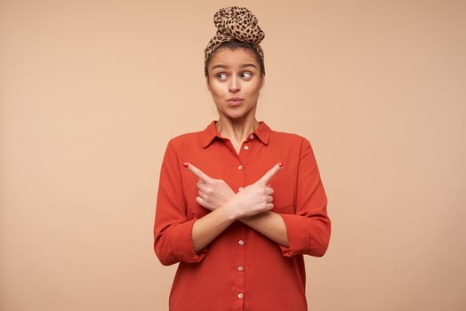 Bewildered young lovely brown haired lady with natural makeup crossing hands with raised forefingers while looking surprisedly aside, posing over beige background photo