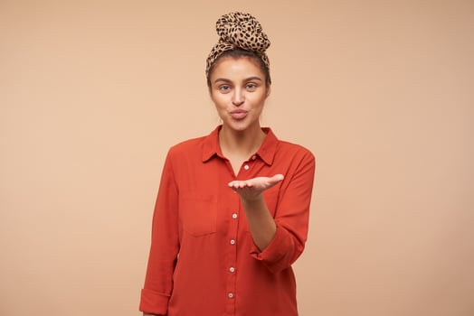 Pleased young attractive green-eyed brunette female pursing her lips while sending air kiss at camera, standing over beige background with raised hand photo