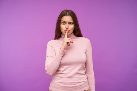 Serious young pretty brown haired woman with natural makeup frowning her eyebrows while looking severely at camera, raising hand with hush gesture while posing over purple background photo