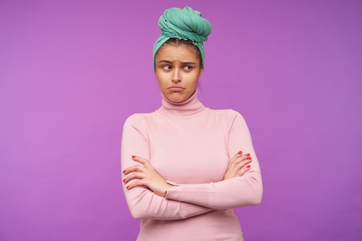 Unhappy young lovely brown haired female with mint headband folding sadly her lips while looking aside and crossing hands on her chest, isolated over purple background photo