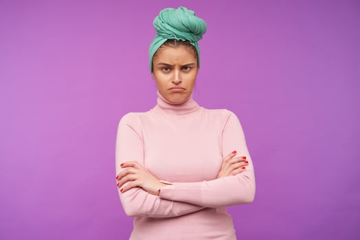 Offended young brown haired female with natural makeup folding hands on her chest while looking sadly at camera, posing over purple background in elegant wear photo