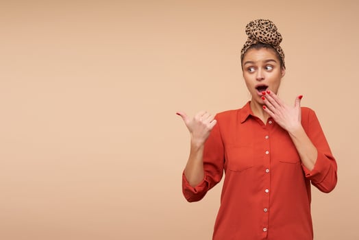 Indoor shot of young elegant brunette female with red manicure covering her mouth with raised hand while looking dazedly aside, isolated over beige background photo