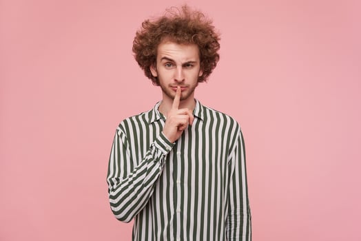 Portrait of lovely young curly redhead male dressed in casual clothes keeping forefinger on his mouth while looking at camera with raised eyebrow, standing against pink background photo