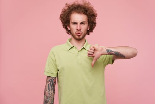 Displeased young curly brunette man with tattoos frowning eyebrows while looking discontentedly at camera while showing with thumb down, isolated over pink background photo
