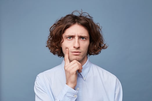 Thoughts and doubts concept. Man with long messy hair thoughtful face, holding fingers on cheek and chin, gazing camera. Guy with untidy hairstyle has doubts, copy space, blue background. photo