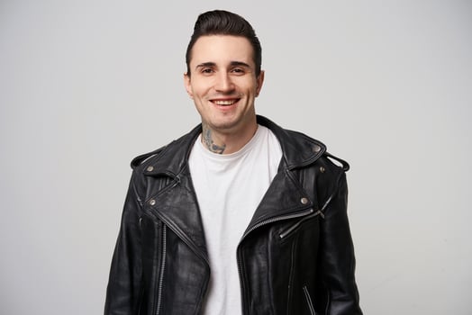 Attractively smiling young rebel, with a stylish hairstyle in briolin, tattoo on the neck, dressed in the style of 50s-60s in black leather jacket and white T-shirt, over white background photo