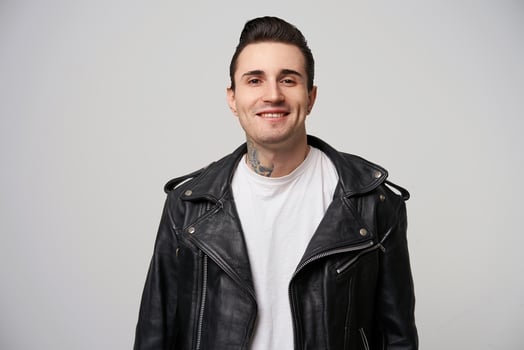 Charmingly, enticing, boldly smiling young rocker, rebel, motto racer with a stylish hairstyle in briolin, tattoo on the neck, dressed in the style of 50s-60s in black leather jacket, white background photo
