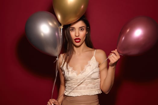 Studio shot of young brown-eyed long haired brunette woman with evening makeup holding air balloons and looking seriously at camera, isolated over burgundy background photo