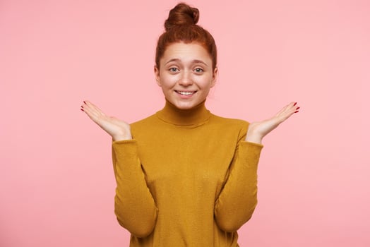 Portrait of attractive, adult girl with ginger hair, freckles and bun. Wearing gold turtleneck sweater and smile. Showing confusion. Watching at the camera isolated over pastel pink background photo