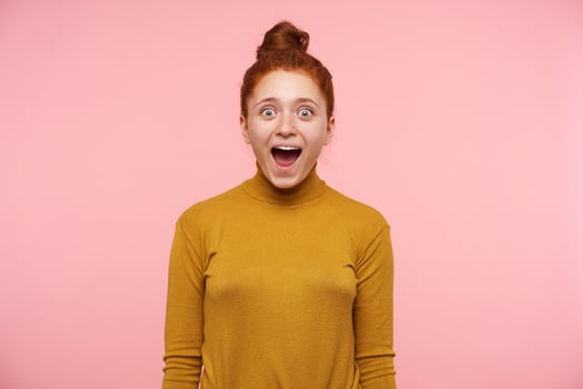 Portrait of attractive, shocked, adult girl with ginger hair, freckles and bun. Wearing gold turtleneck sweater. Emotion concept. Watching at the camera isolated over pastel pink background photo