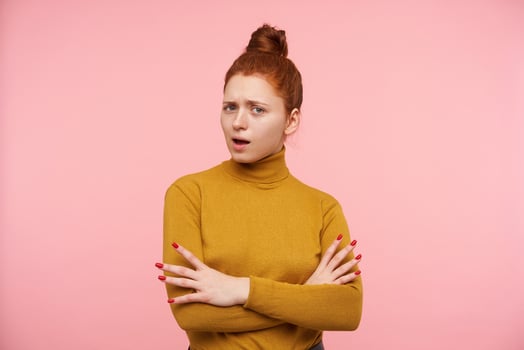 Portrait of attractive, adult girl with ginger hair, freckles and bun. Wearing gold turtleneck sweater and holding hands crossed. Watching suspicious at the camera isolated over pastel pink background photo