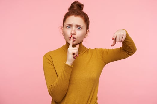 Severe young redhead pretty lady with casual hairstyle frowning her eyebrows while looking seriously at camera and raising hand with hush gesture, isolated over pink background photo